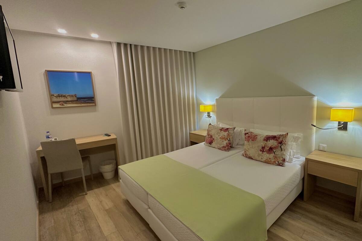 Ein Hotelzimmer mit einem Bett, Nachttischen, einem Schreibtisch, einem Sessel, einer Lampe und einem Bild an der Wand.