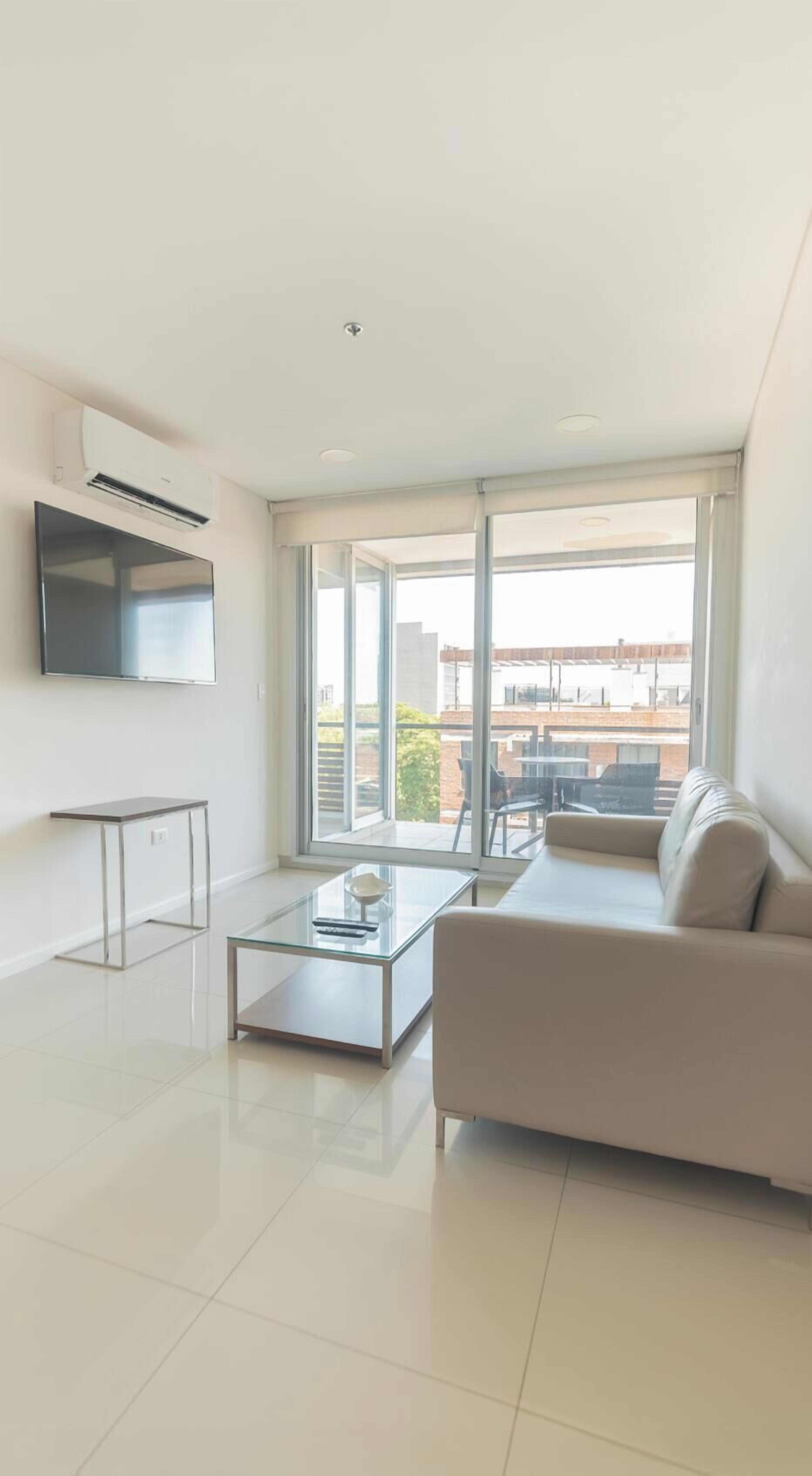 Una sala de estar moderna con un sofá, una mesa de centro de vidrio, un televisor montado en la pared y ventanas amplias con vistas al exterior.