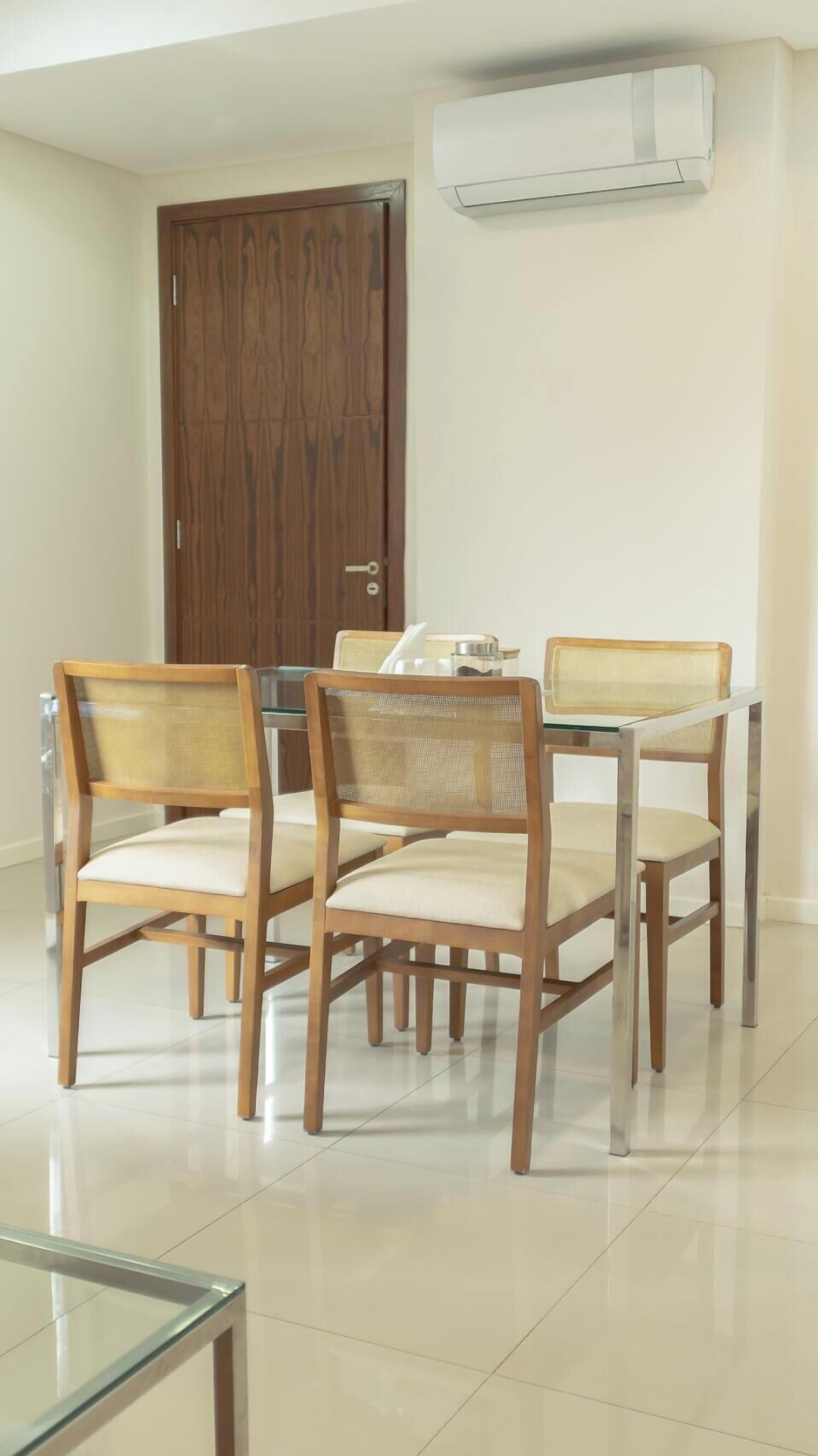 A dining area with wooden chairs and a glass table, a wooden door, and an air conditioning unit on the wall.