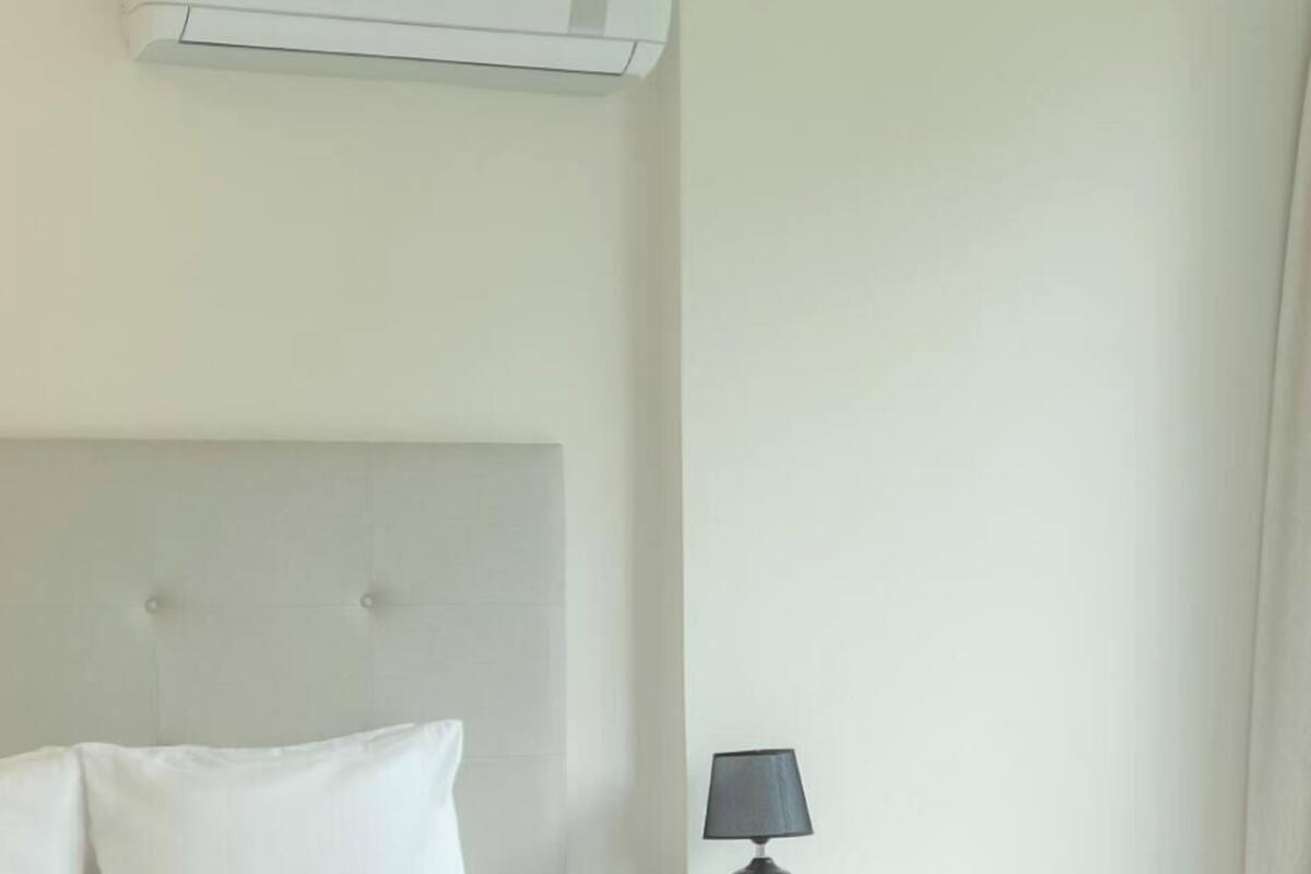 A bed with white bedding, a bedside lamp, and a wall-mounted air conditioner.