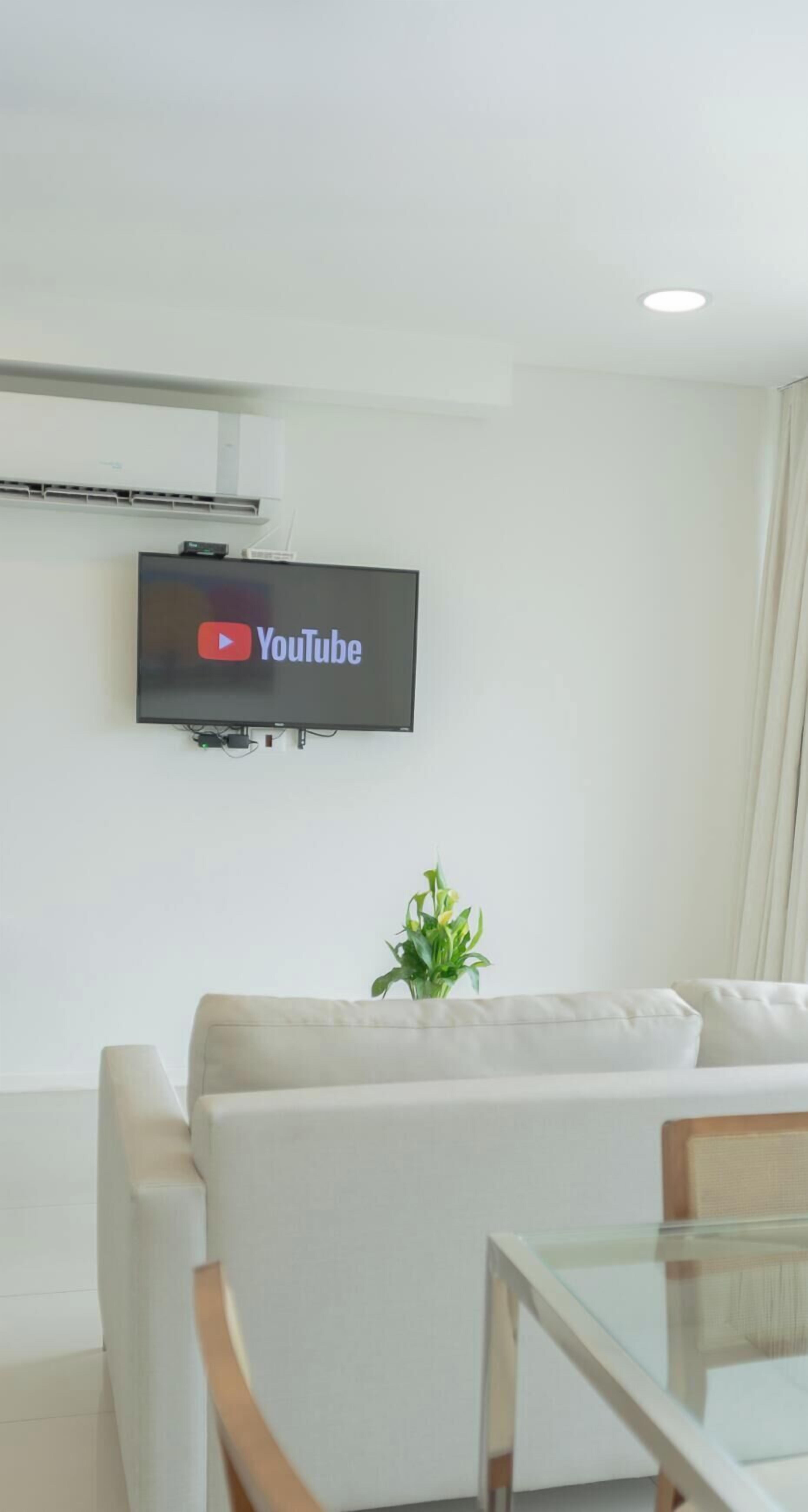 A living room with a flat-screen TV mounted on the wall displaying the YouTube logo, a white sofa, a glass table, and a small plant.