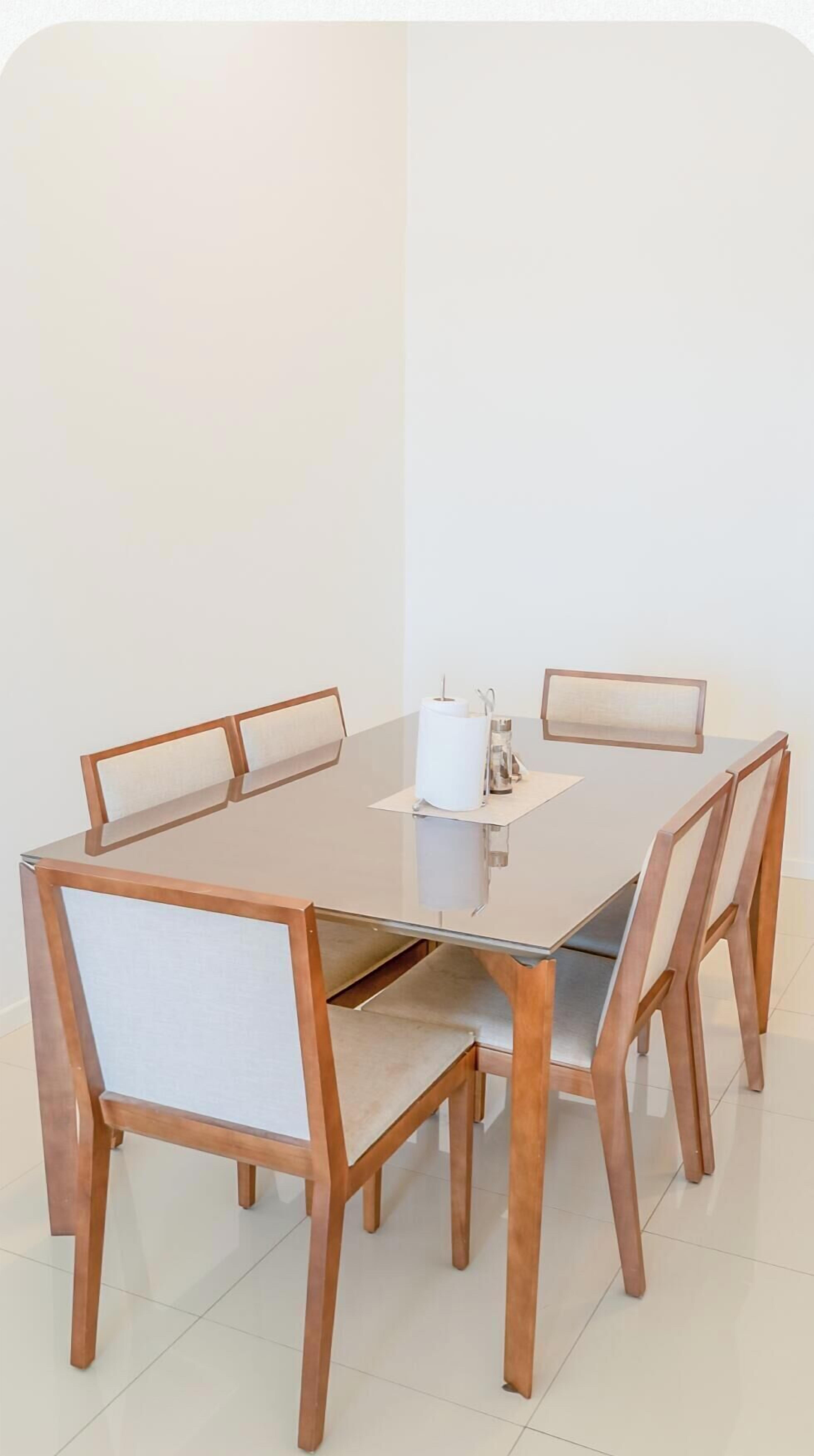 Una mesa rectangular con cubierta de vidrio, sillas de madera, un rollo de toallas de papel y una jarra de vidrio sobre la mesa.