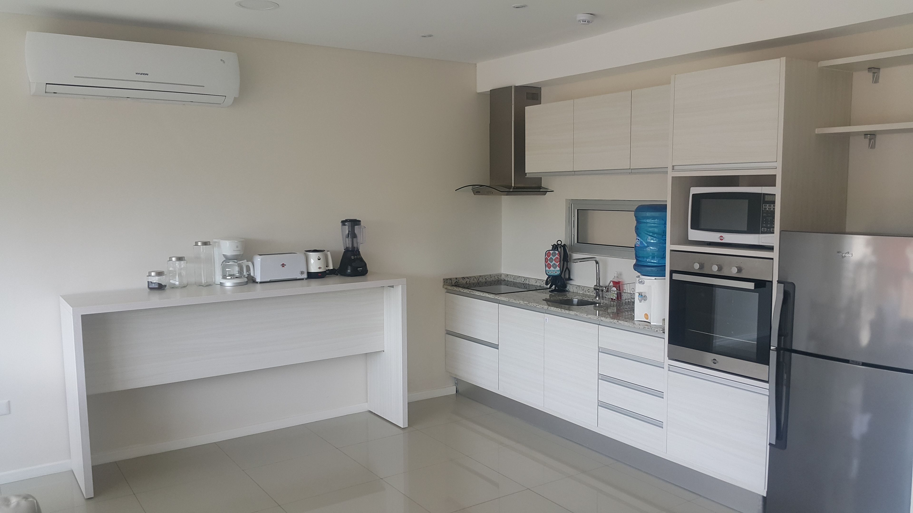 Una cocina moderna con armarios blancos, microondas integrado, refrigerador de acero inoxidable y cafetera sobre la encimera.