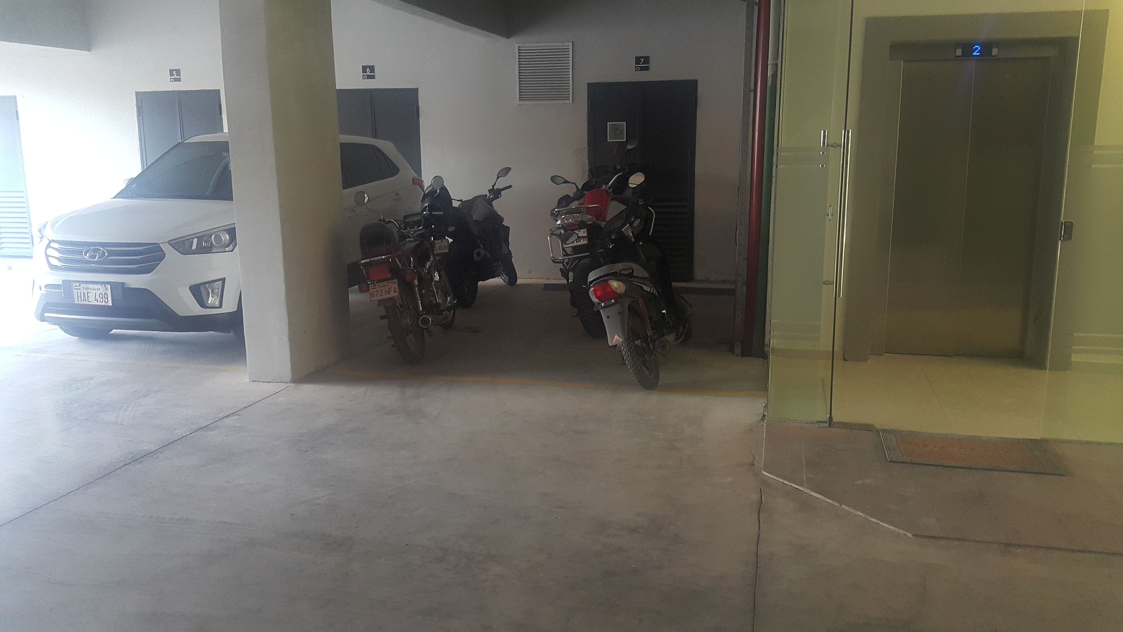 Un estacionamiento subterráneo con varias motocicletas y un coche blanco. Se observan plazas de aparcamiento numeradas y la puerta de un ascensor de cristal.