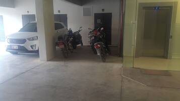 Un estacionamiento subterráneo con varias motocicletas y un coche blanco. Se observan plazas de aparcamiento numeradas y la puerta de un ascensor de cristal.