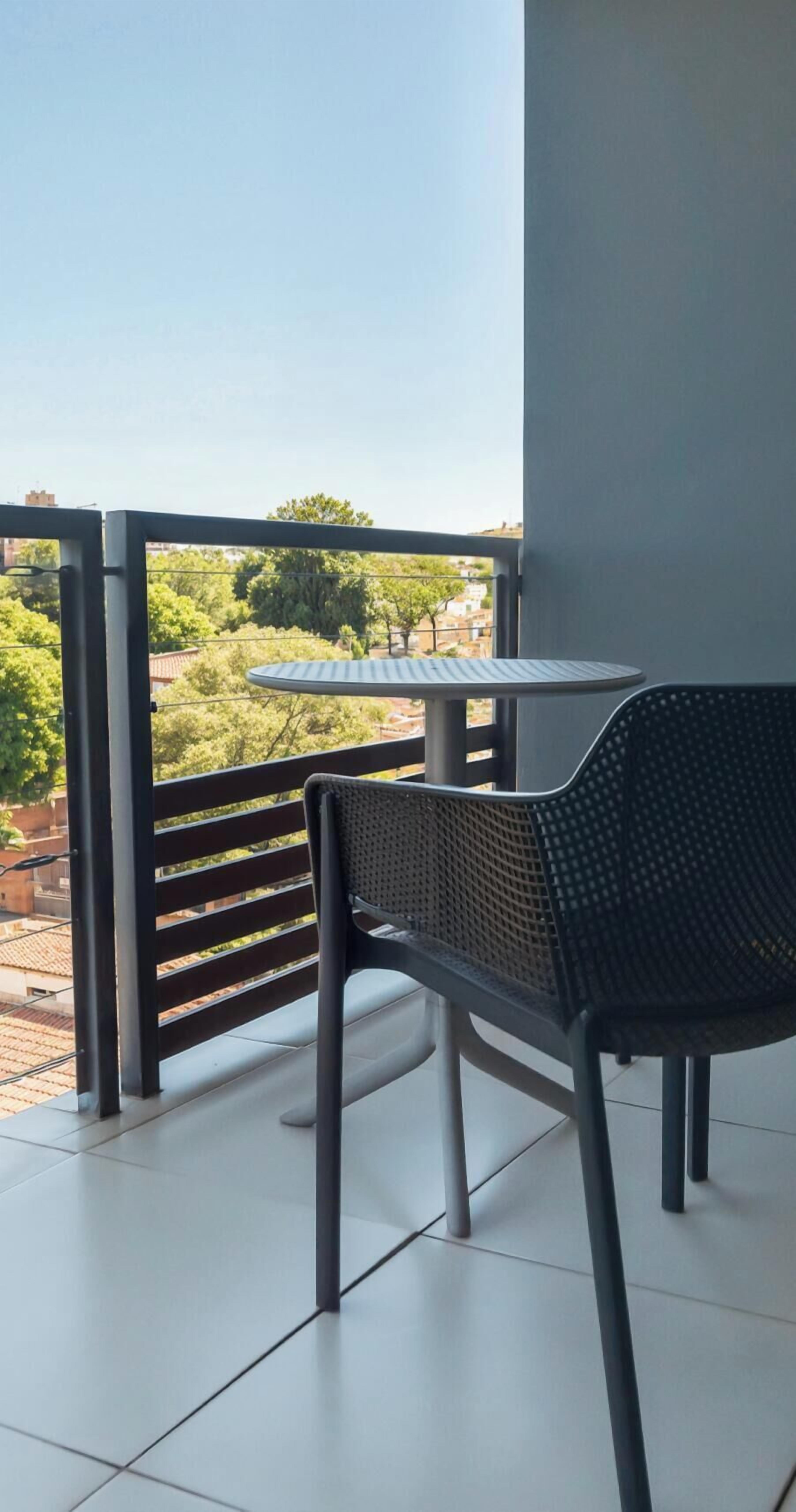 A balcony with a table and chairs, a view of greenery and buildings, and a clear sky.