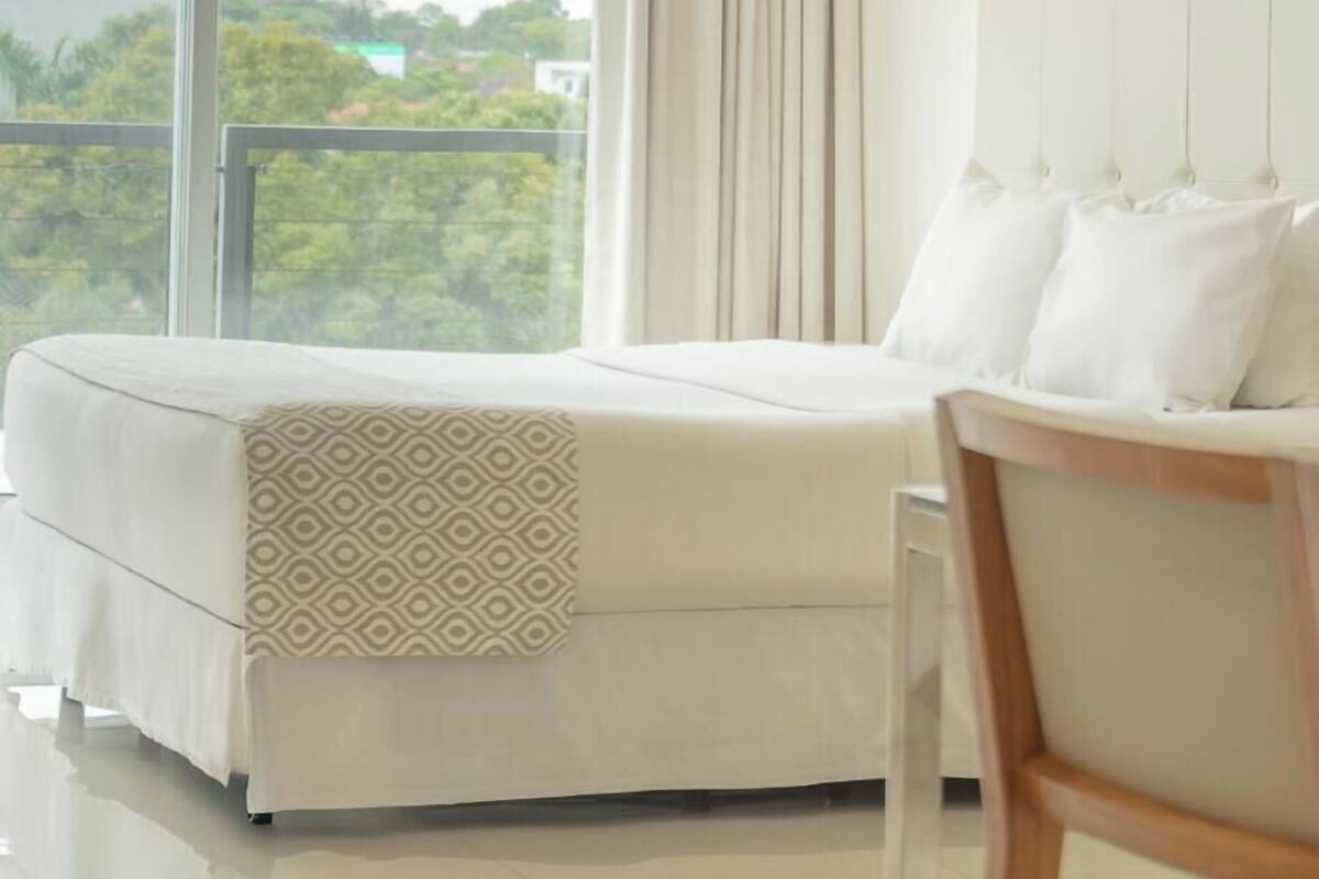 A bedroom with a bed, a chair, a large window with curtains, and a view of buildings and trees outside.