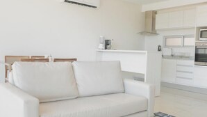 Ein modernes Wohnzimmer mit einem weißen Sofa, einem Glastisch und einer Küche im Hintergrund.