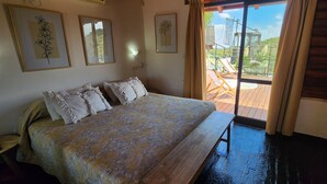 Une chambre à coucher avec un lit, des oreillers, un banc et une vue sur un balcon aménagé avec du mobilier d’extérieur.