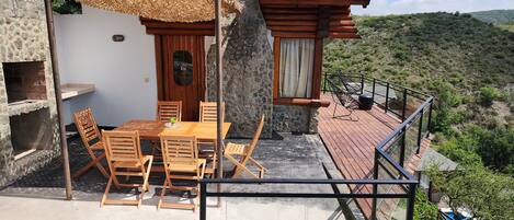 A wooden deck with a table and chairs, a stone wall, and a view of the surrounding hills.