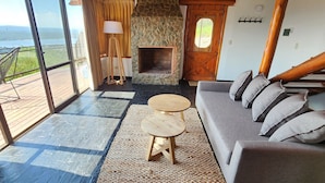 Une pièce spacieuse avec une grande fenêtre, une cheminée en pierre, un canapé gris et une table en bois.