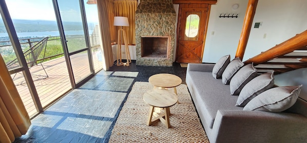 Una habitación amplia con un gran ventanal, una chimenea de piedra, un sofá gris y una mesa de madera.
