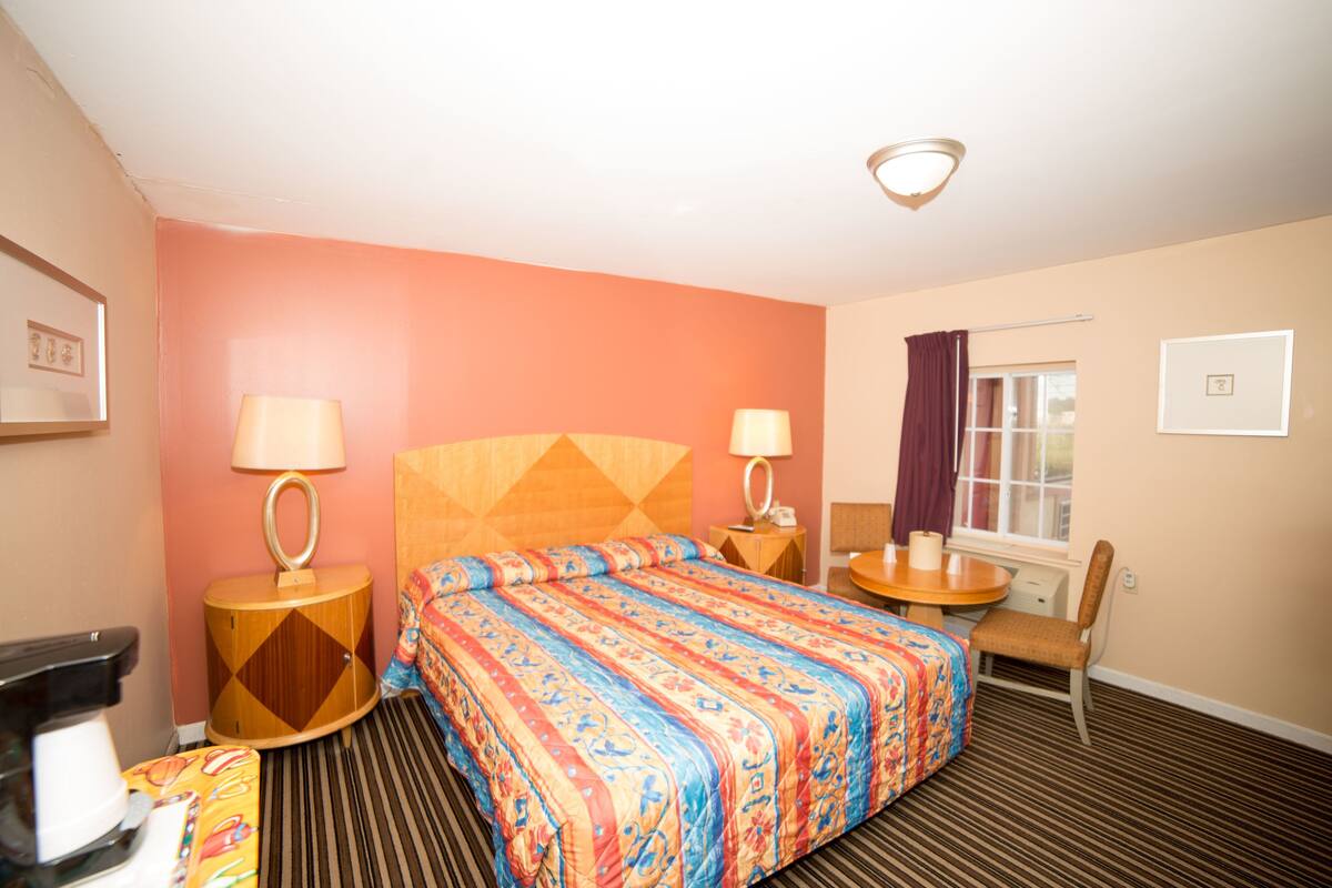 A hotel room with a large bed, two bedside tables with lamps, a small round table with a chair, a window with curtains, and a wall-mounted picture frame.