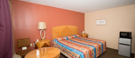 A hotel room with a bed, a round table, a chair, a desk, a lamp, a microwave, and a refrigerator.