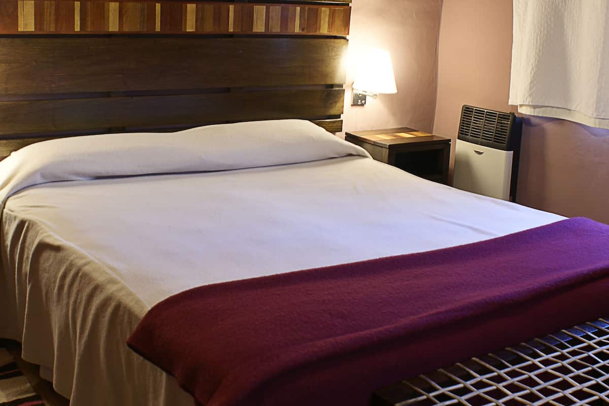 A neatly made bed with white linens and a red throw blanket, a bedside table with a lamp, and a small heater on the wall.