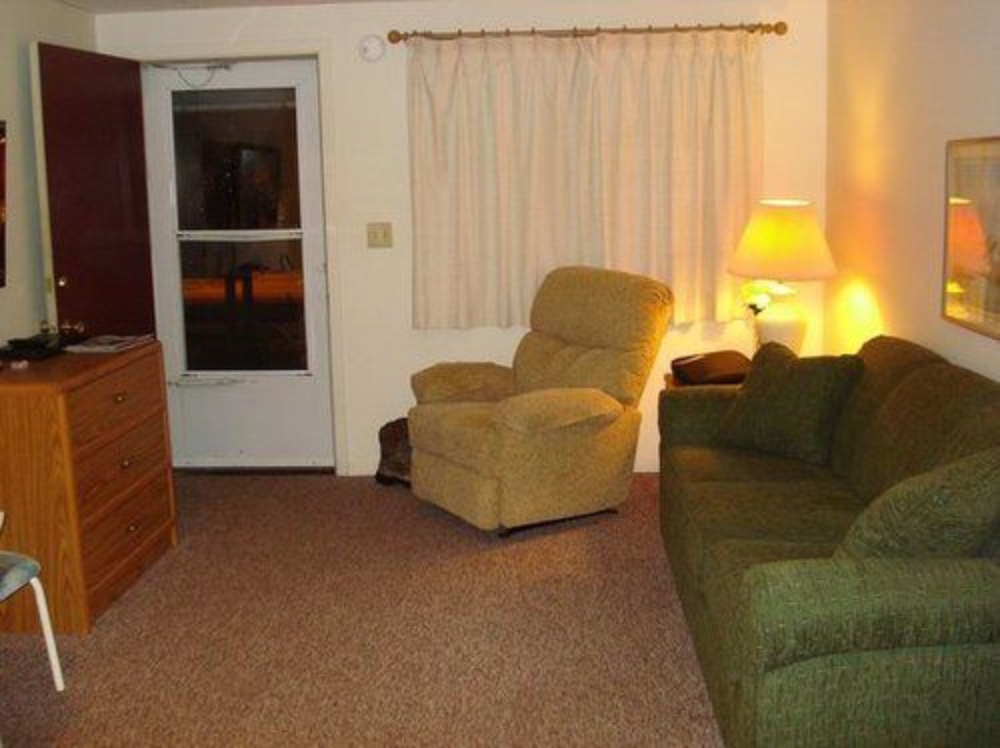 A room with a green sofa, a brown armchair, a wooden dresser, and a lamp on a side table.