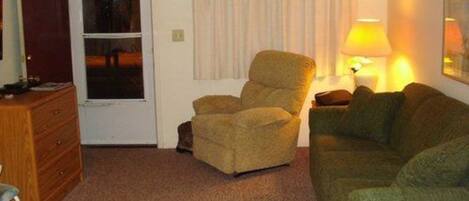 A room with a green sofa, a brown armchair, a wooden dresser, and a lamp on a side table.