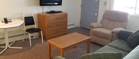 A room with a television, a wooden dresser, a green sofa, a beige armchair, and a small table.