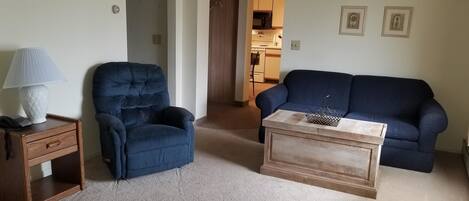 A living room with a blue armchair, a blue sofa, a wooden coffee table, and a lamp on a nightstand.