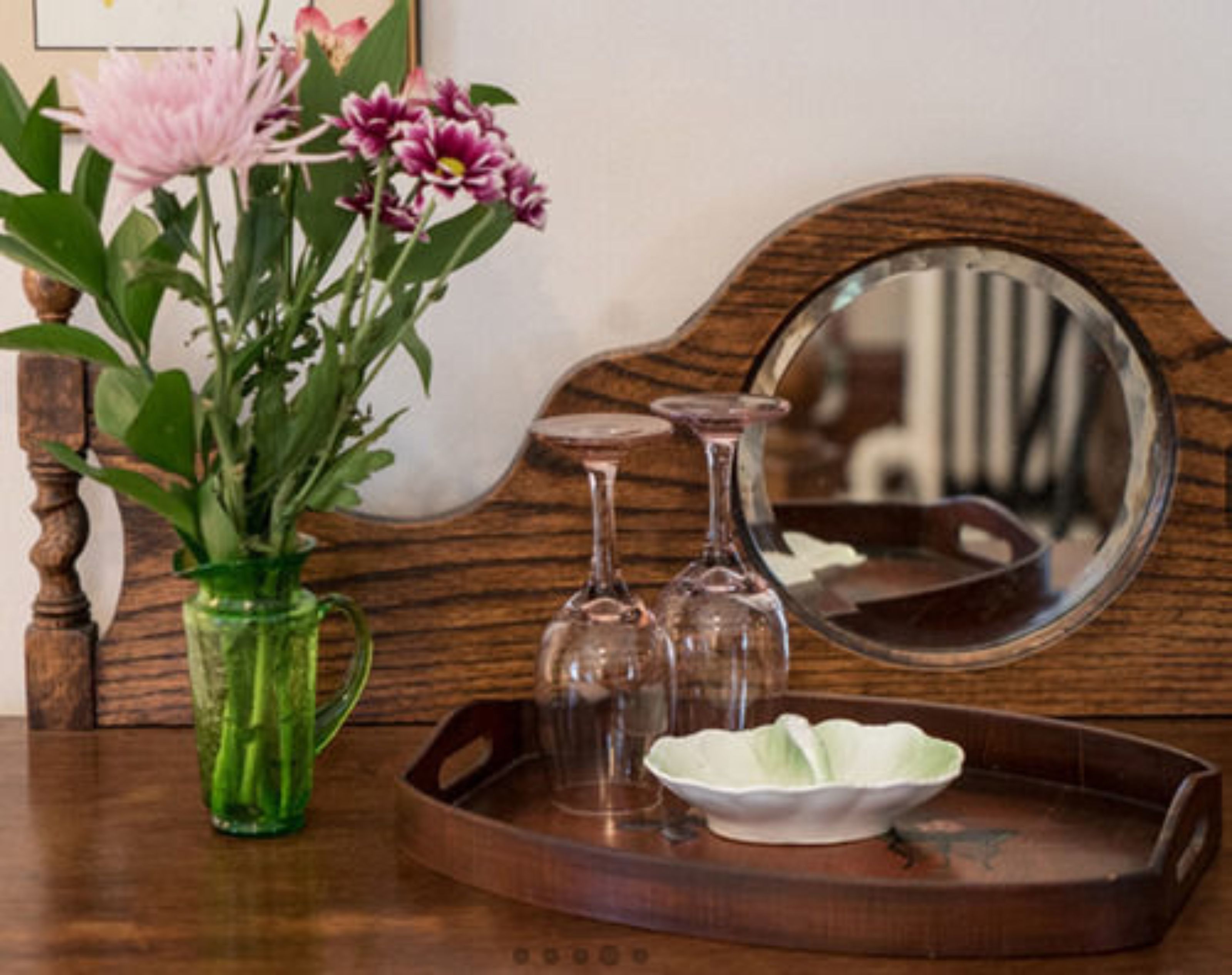 A wooden side table with a green vase containing flowers, a tray with a white bowl, and a mirror.