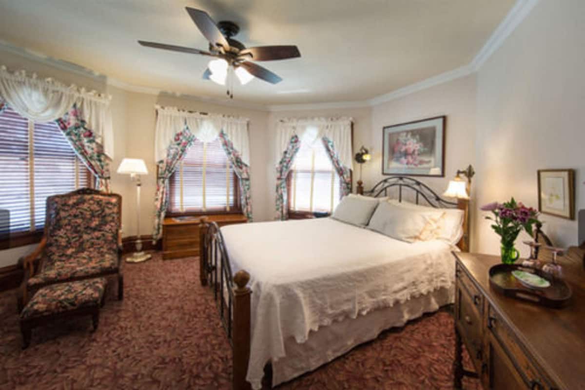 Une chambre à coucher comprenant un lit, une chaise, une commode, un ventilateur de plafond et des fenêtres avec des rideaux.