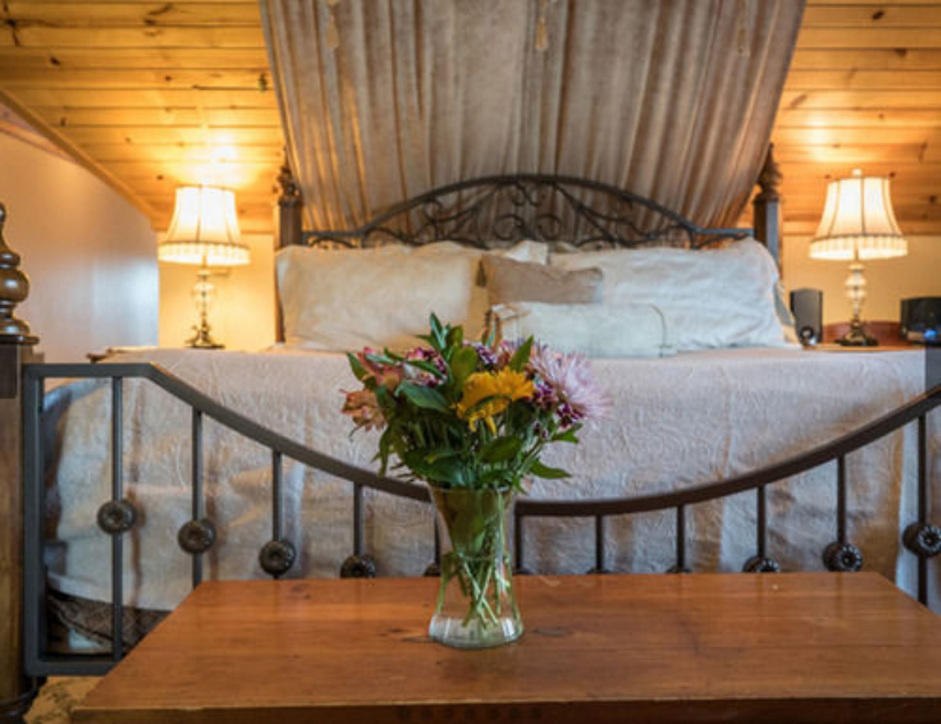 A bedroom with a wooden headboard, a bed with white linens, two bedside lamps, and a vase with flowers on a wooden table.