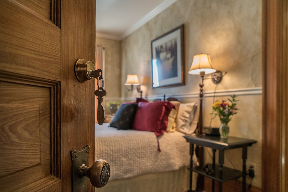 Une chambre d’hôtel avec une porte en bois, un lit recouvert d’une couette beige, deux lampes de chevet, un tableau encadré au mur et une petite table sur laquelle est posé un vase de fleurs.