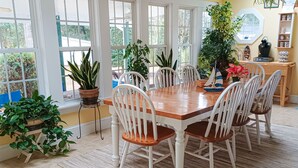 A dining area with a wooden table and white chairs, surrounded by large windows and potted plants. - The Inn on Bath Creek (Bath)