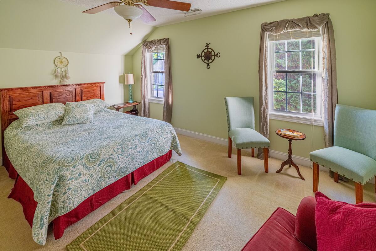 Quarto com uma cama grande, um tapete verde, uma cadeira, uma mesa pequena e uma janela com cortinas.