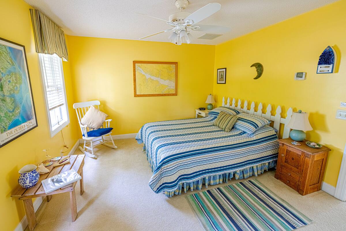 Quarto com cama, cadeira, mesa de cabeceira, luminária, mapa e ventilador de teto.