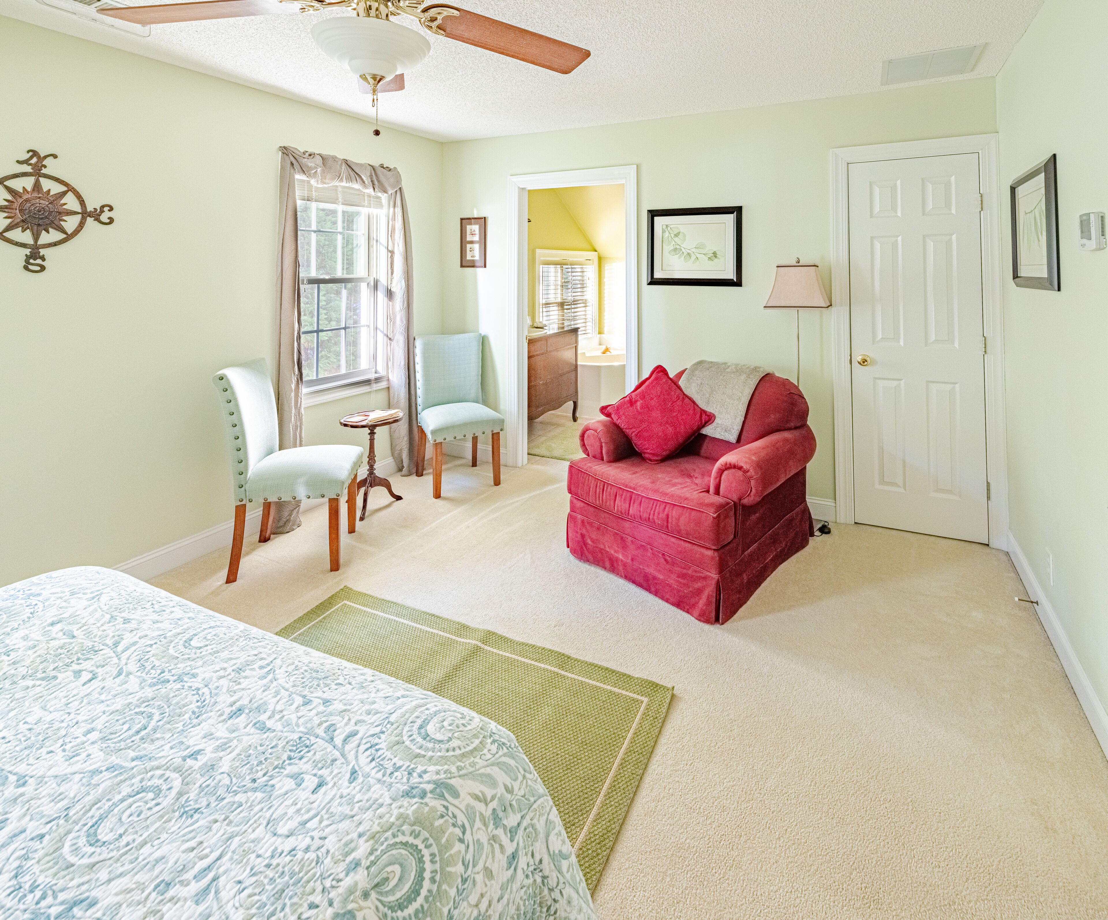 A room with a bed, a red armchair, a blue armchair, a wooden chair, a small table, a lamp, and a door.