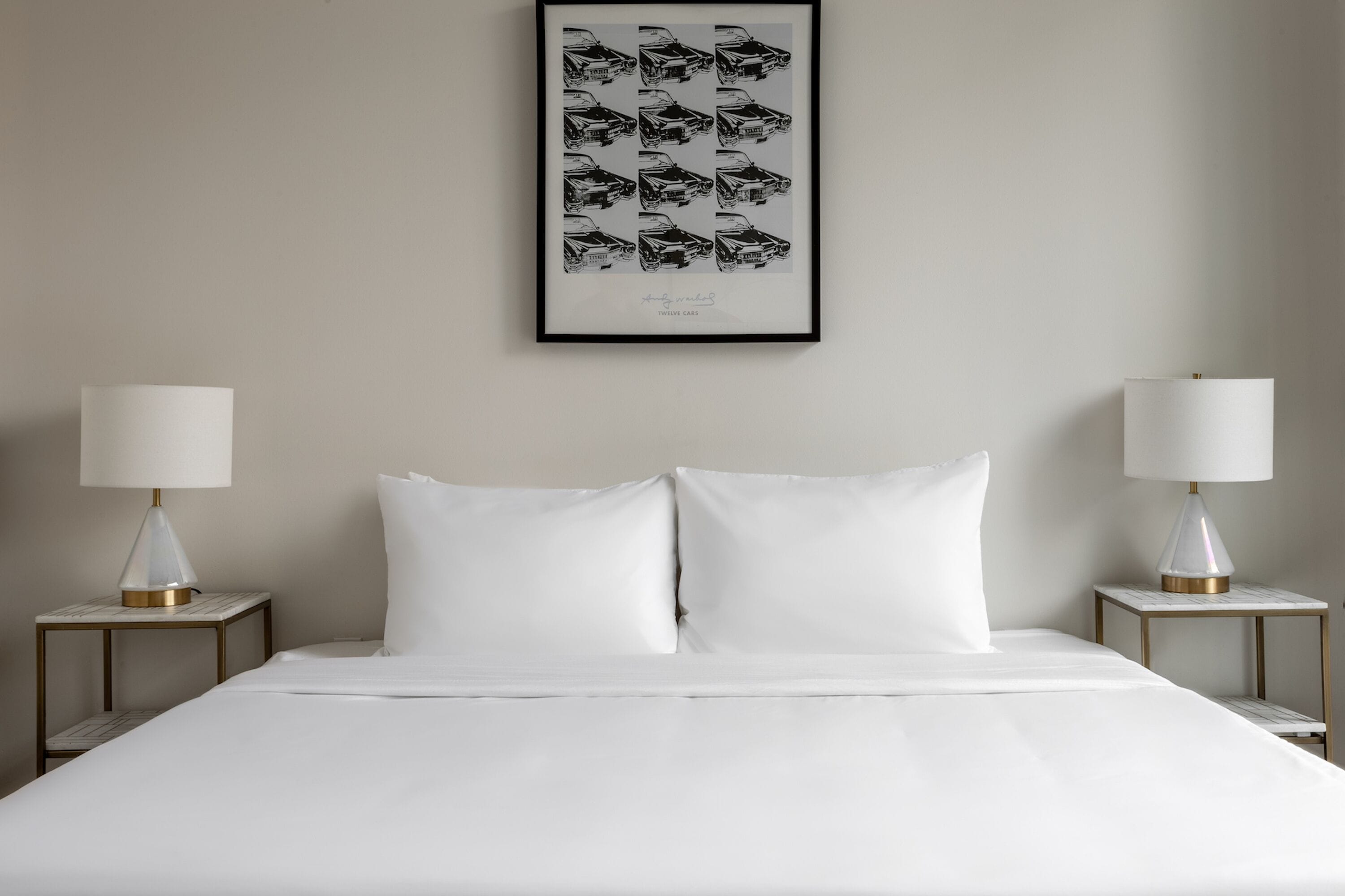 A neatly made bed with white linens, two bedside tables with lamps, and a framed picture of cars on the wall.