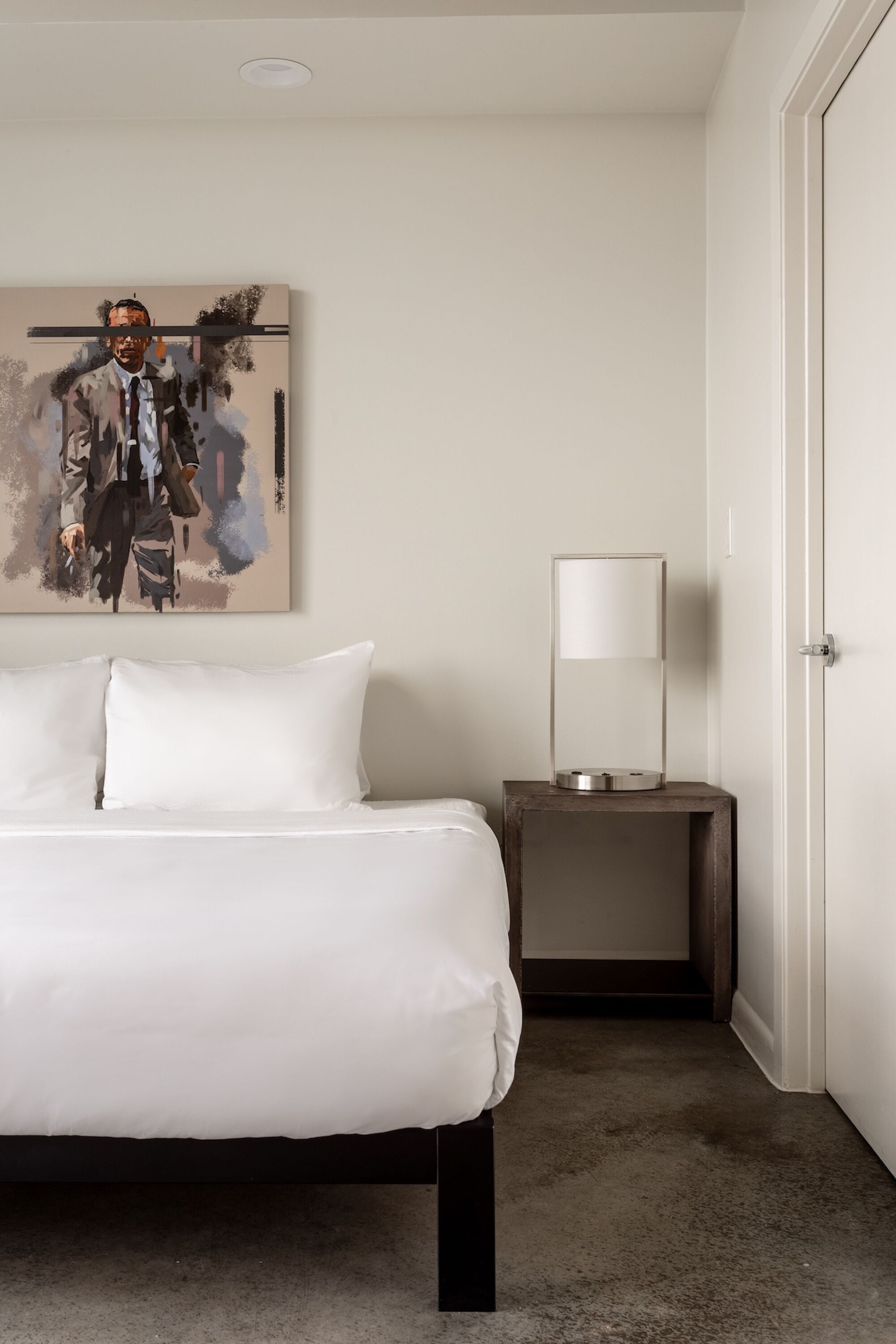 A hotel room with a bed, a bedside table with a lamp, and a painting on the wall.