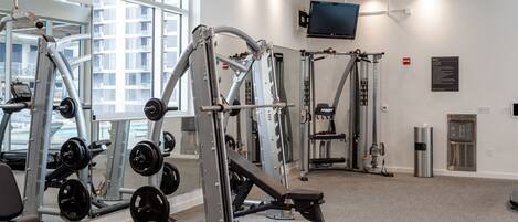 A gym with various exercise machines, a large mirror, a television on the wall, and a view of the outside through large windows.