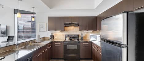 A modern kitchen with dark wooden cabinets, stainless steel appliances, a large window, and a concrete ceiling.