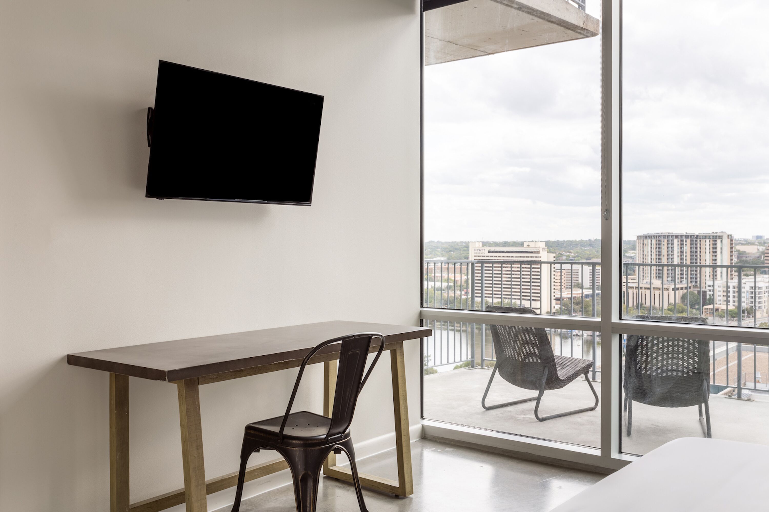 A modern hotel room with a large window, a flat-screen TV mounted on the wall, a wooden desk, and two chairs.