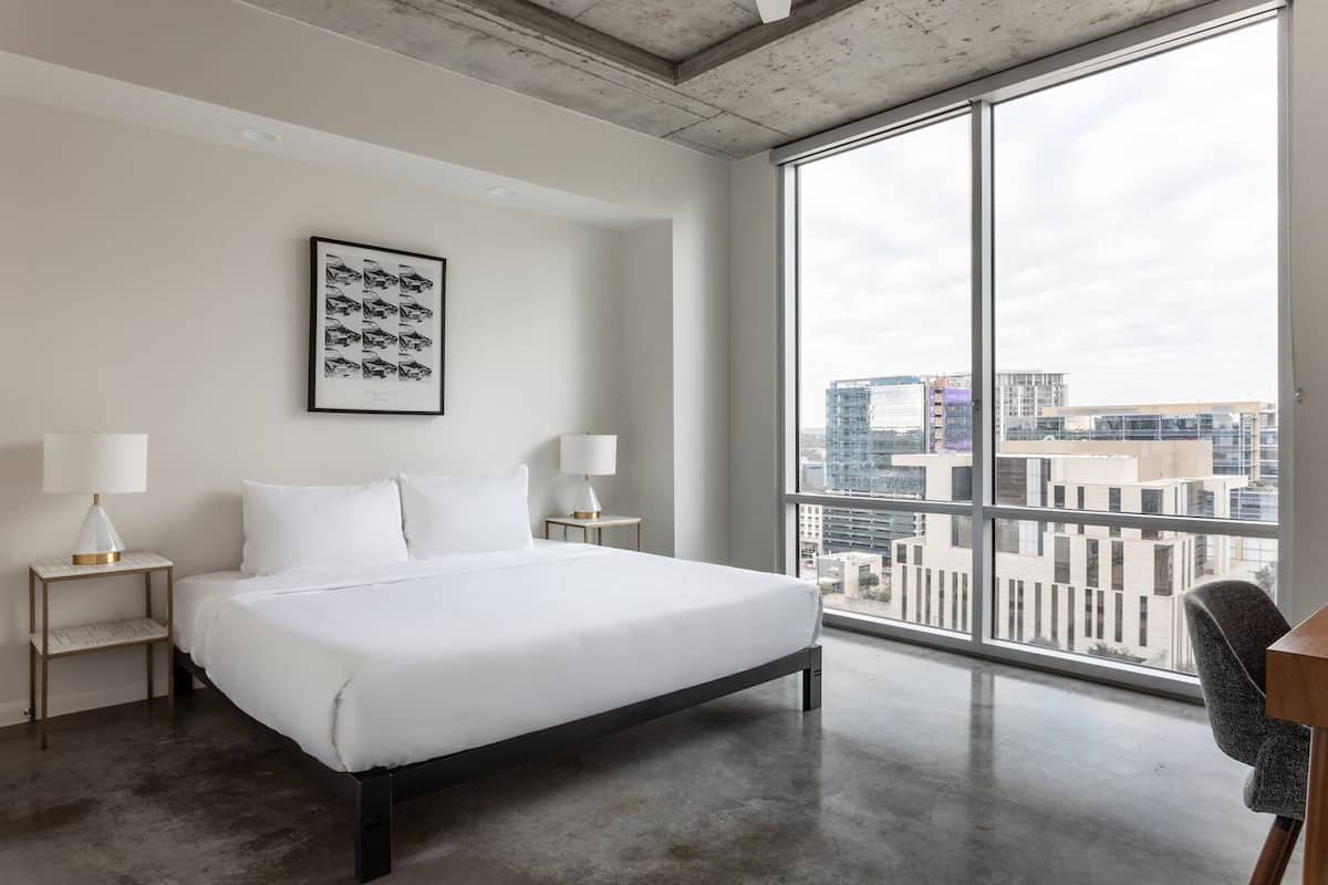 Ein modernes Hotelzimmer mit einem großen Bett, zwei Nachttischen mit Lampen, Blick auf die Stadt durch raumhohe Fenster und einem gerahmten Bild an der Wand.