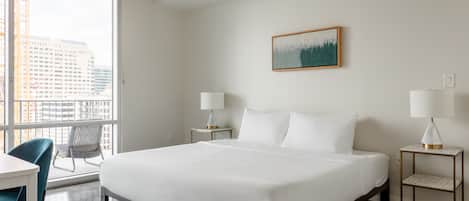 Ett hotellrum med en stor säng, två sängbord med lampor, en stol och utsikt över byggnader genom ett stort fönster.