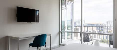 A hotel room with a large window, a flat-screen TV, a white desk, a blue chair, and a small white table.