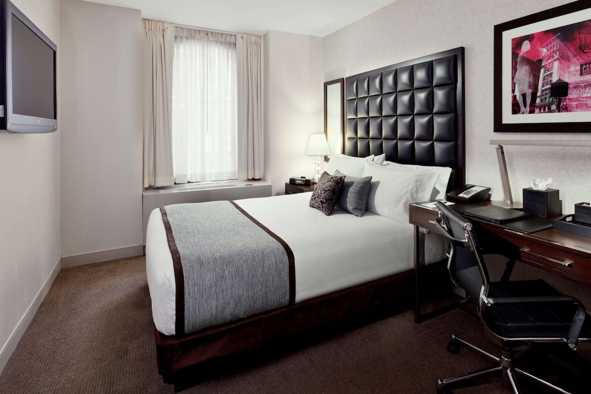 Une chambre d’hôtel avec un grand lit, un bureau avec une chaise, une fenêtre avec des rideaux et un tableau encadré au mur.