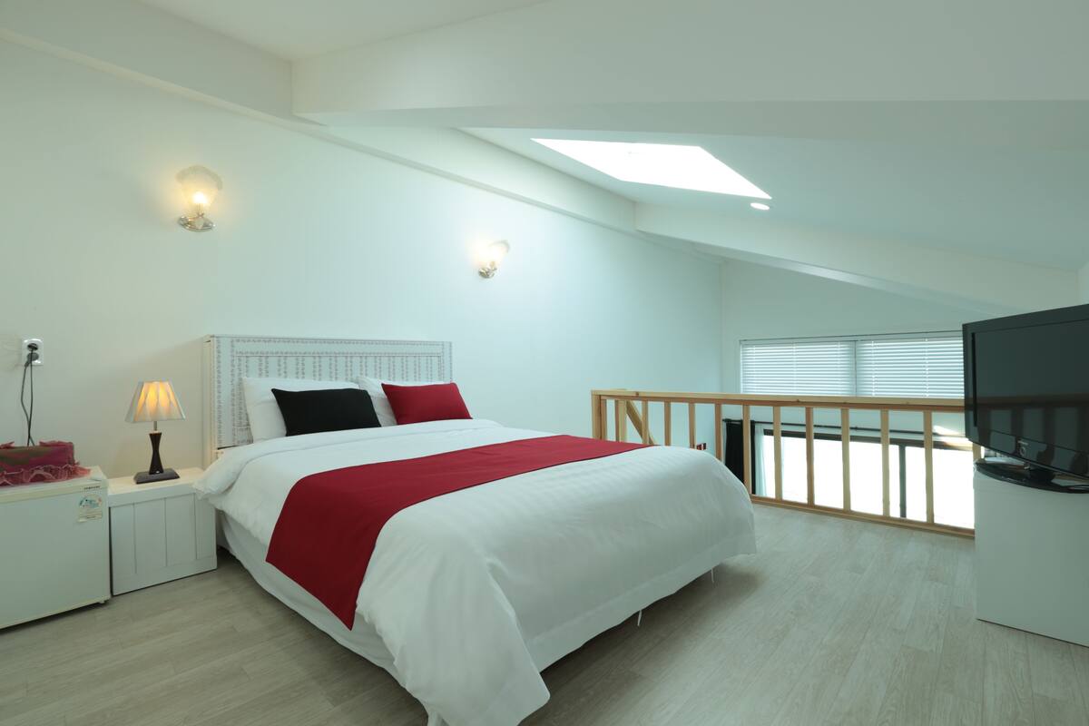 A modern bedroom with a skylight, a bed with white and red bedding, a nightstand with a lamp, and a TV on a stand.