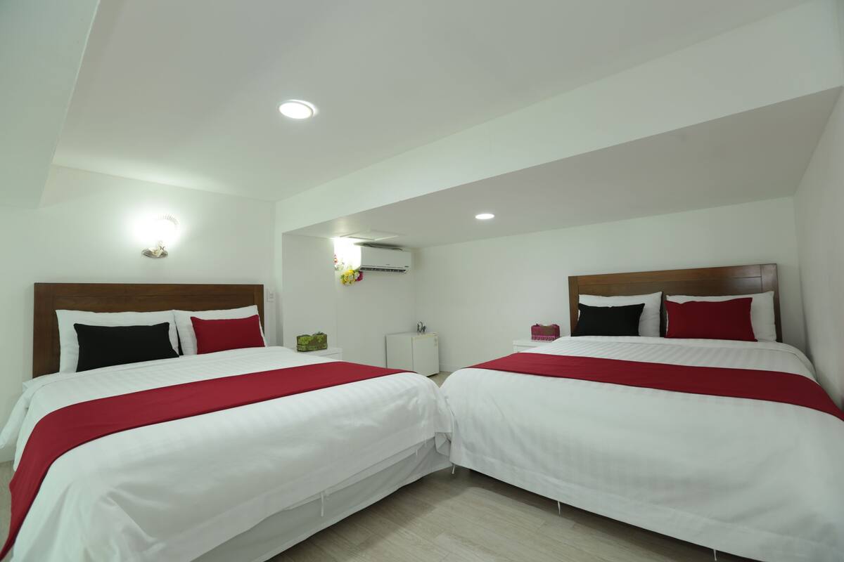 A hotel room with two beds, white bedding, red accents, a wooden headboard, a nightstand, an air conditioning unit, and a wall-mounted light fixture.