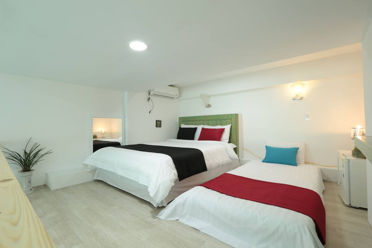 A hotel room with two beds, a bedside table, a lamp, a potted plant, and a wall-mounted air conditioner.