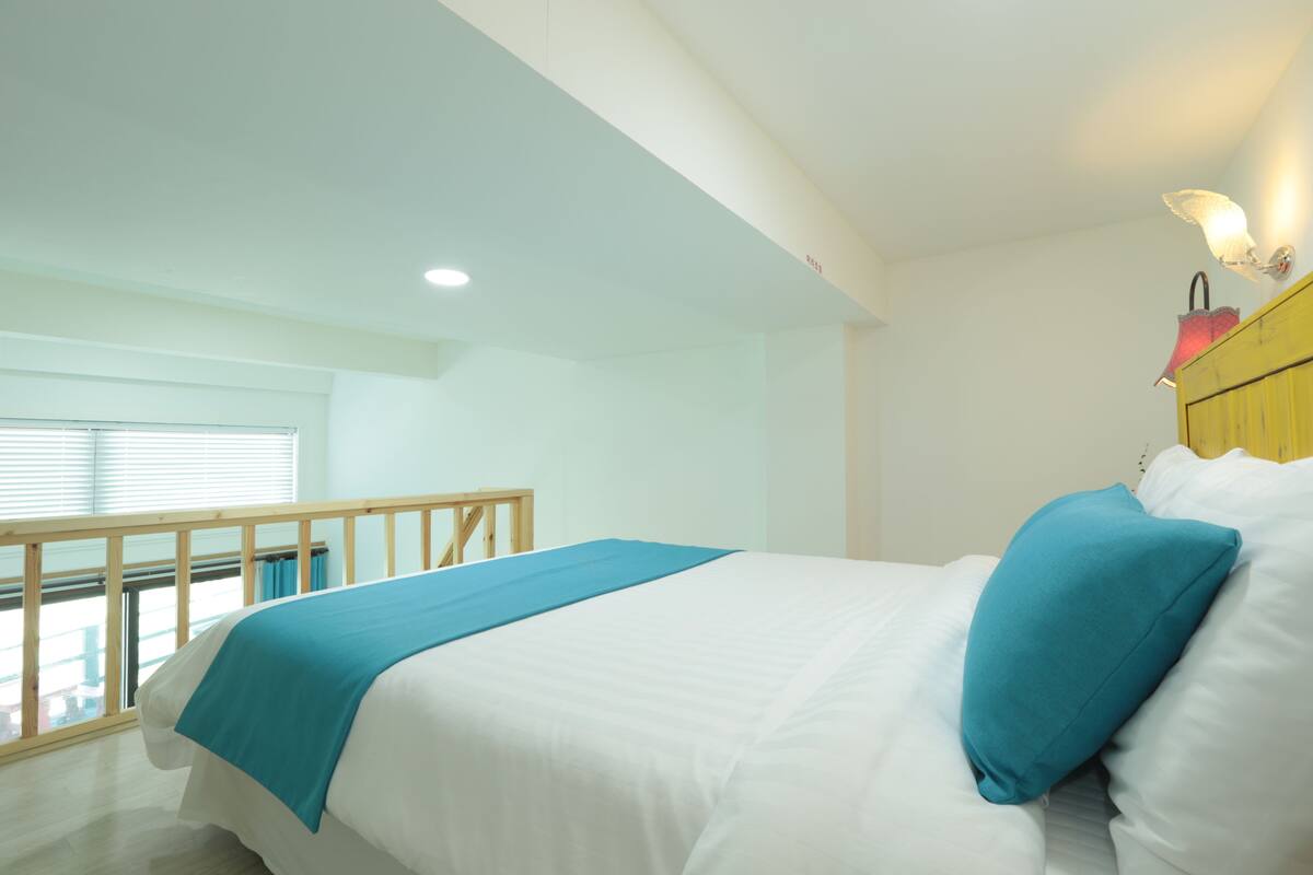 A bedroom with a bed, a blue throw blanket, white pillows, a wooden headboard, and a window with blinds.