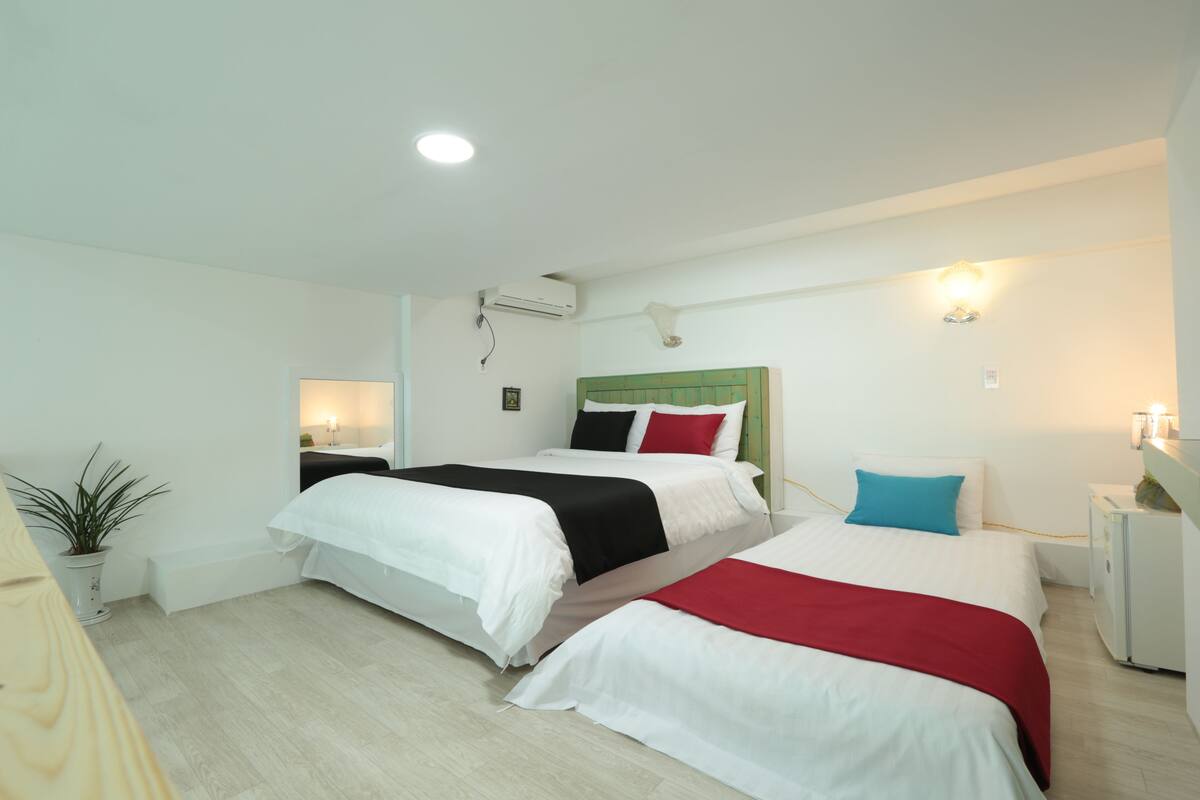 A hotel room with two beds, a bedside table, a lamp, a potted plant, and a wall-mounted air conditioner.