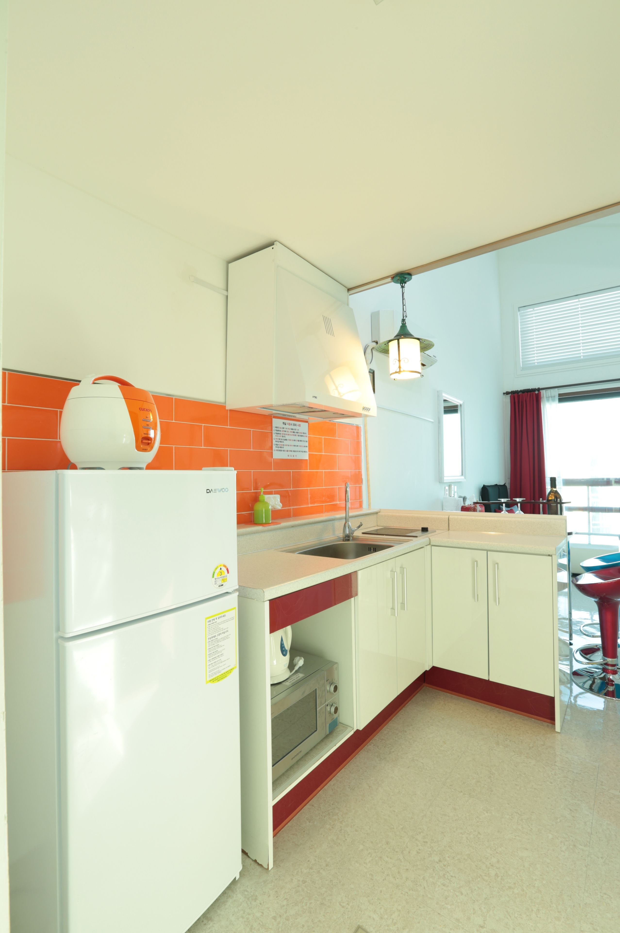 Une cuisine avec un réfrigérateur blanc, un évier et une cuisinière. Il y a une fenêtre avec un rideau rouge et un luminaire suspendu.