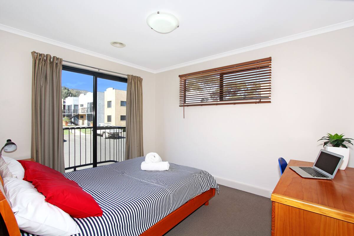 A hotel room with a bed, a desk with a laptop, a window with blinds, and a view of buildings outside.