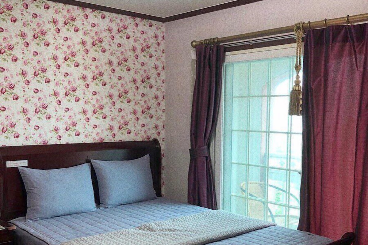 A bedroom with a floral wallpaper, a bed with a quilted cover, and a window with purple curtains.