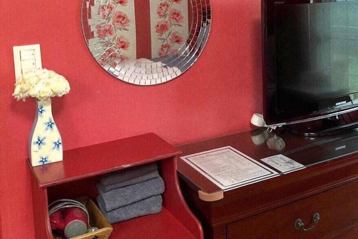 A room with a red wall, a round mirror with floral design, a television, a wooden dresser with a mirror, and a vase with flowers.