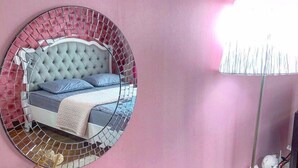 A bedroom with a round mirror, a bed with a tufted headboard, a bedside table with a vase, and a lamp.